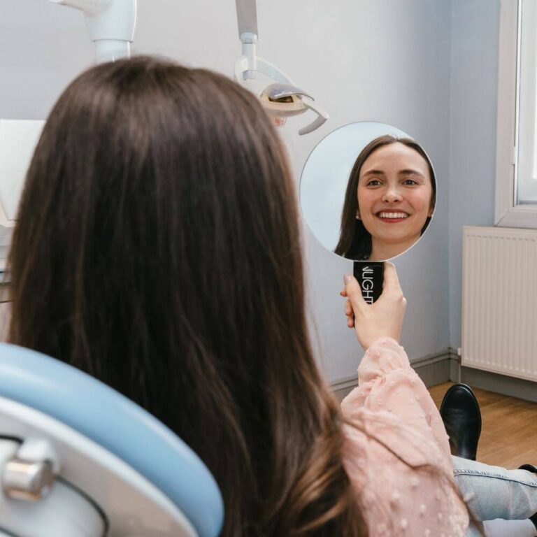 Lady sitting on dental chair holding mirror and smiling at her new white smile after teeth whitening treatment at The Town House.